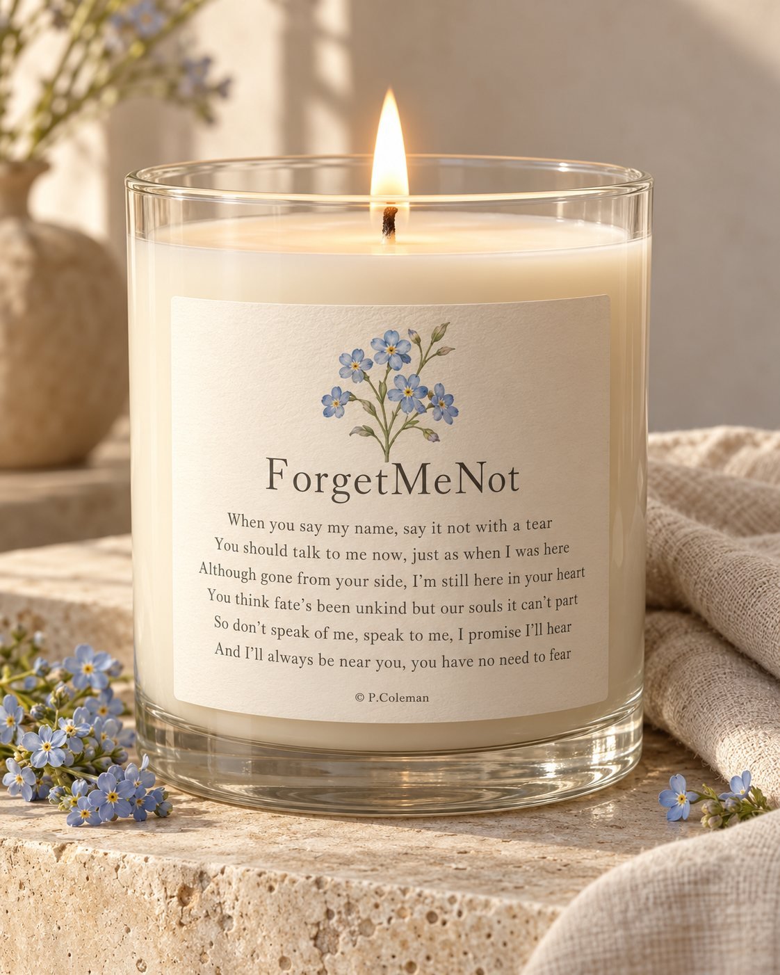 The ForgetMeNot candle, lit, in a clear glass jar with the full verse printed on a cream label beneath a spray of forget-me-nots, © P. Coleman